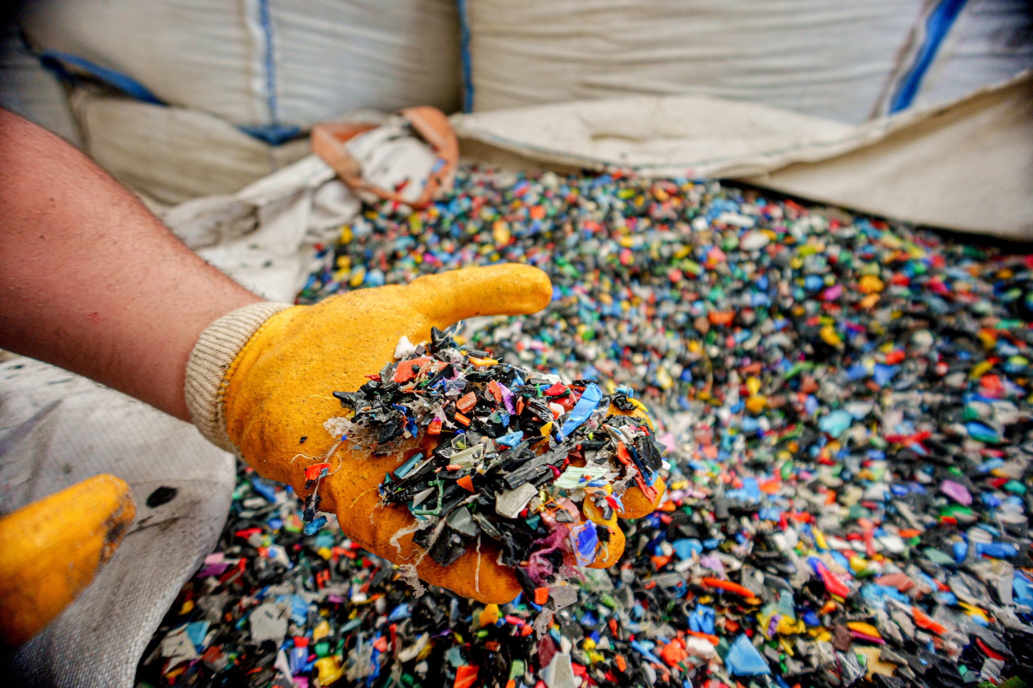 Close-up image of a worker wearing yellow protective gloves holding a handful of multi color recycled plastic pellets. The granules are used as raw materials in plastic manufacturing and injection molding processes. The image represents sustainability, recycling, and industrial production.