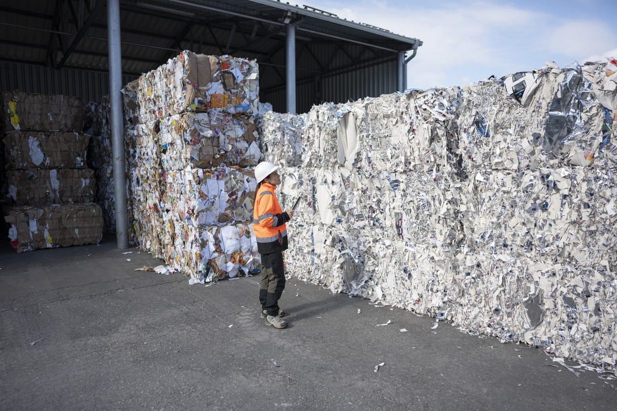 Female worker in high-visibility gear & hardhat uses tablet to inspect massive bales of compacted recycling material outdoors at a waste management facility.