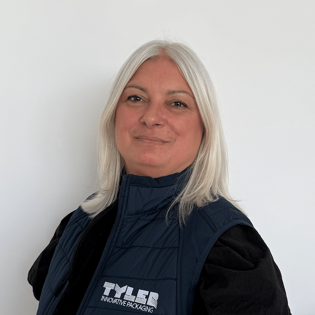 Headshot photograph of Fiona, showing her head and shoulders. She is standing sideways but looking forward. She has short white hair and is wearing a gilet with the "TYLER" logo on it. She's infront of a white background.