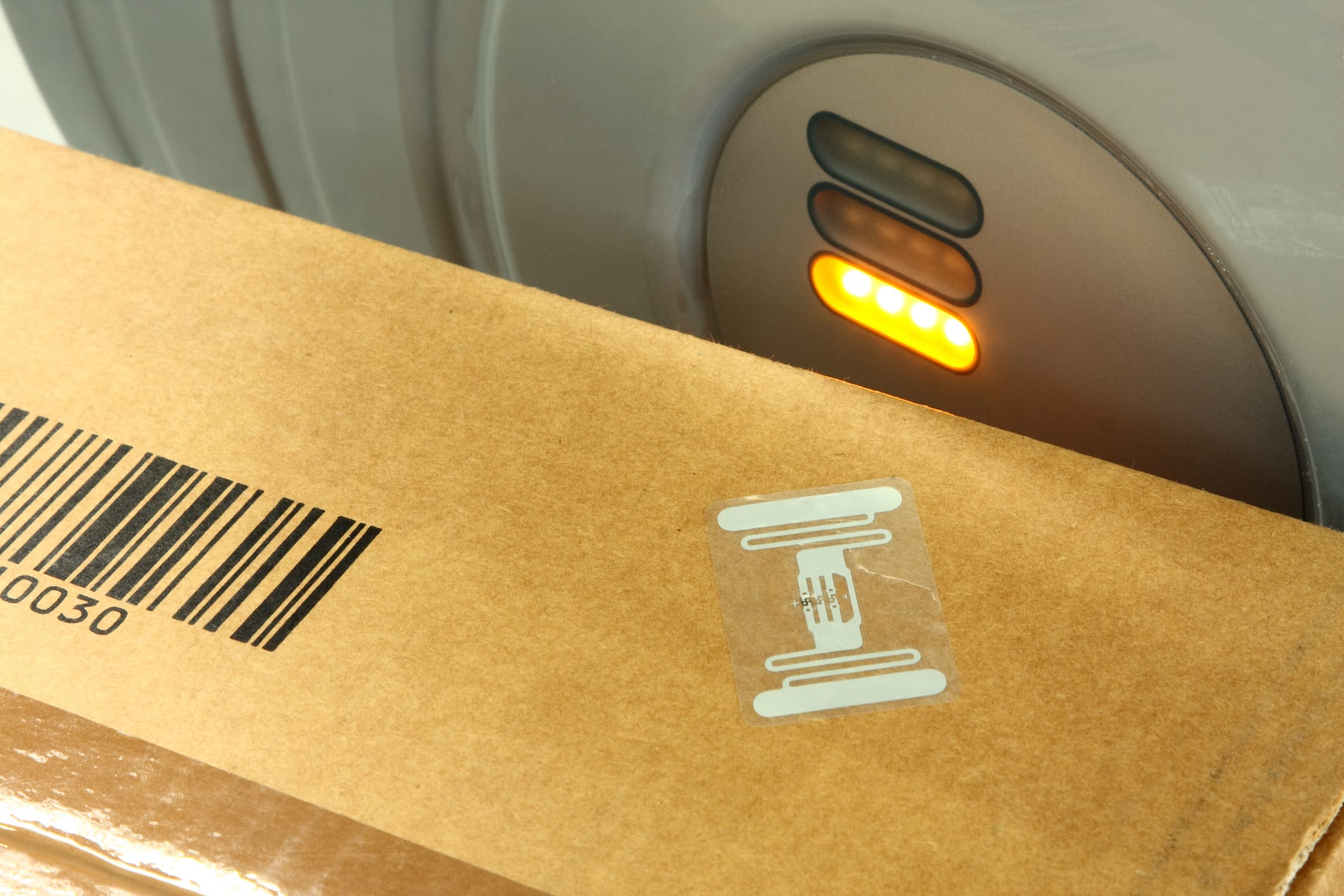 A cardboard box with a barcode and RFID sticker is placed next to an RFID antenna machine with three indicator lights, only one of which is lit in yellow.