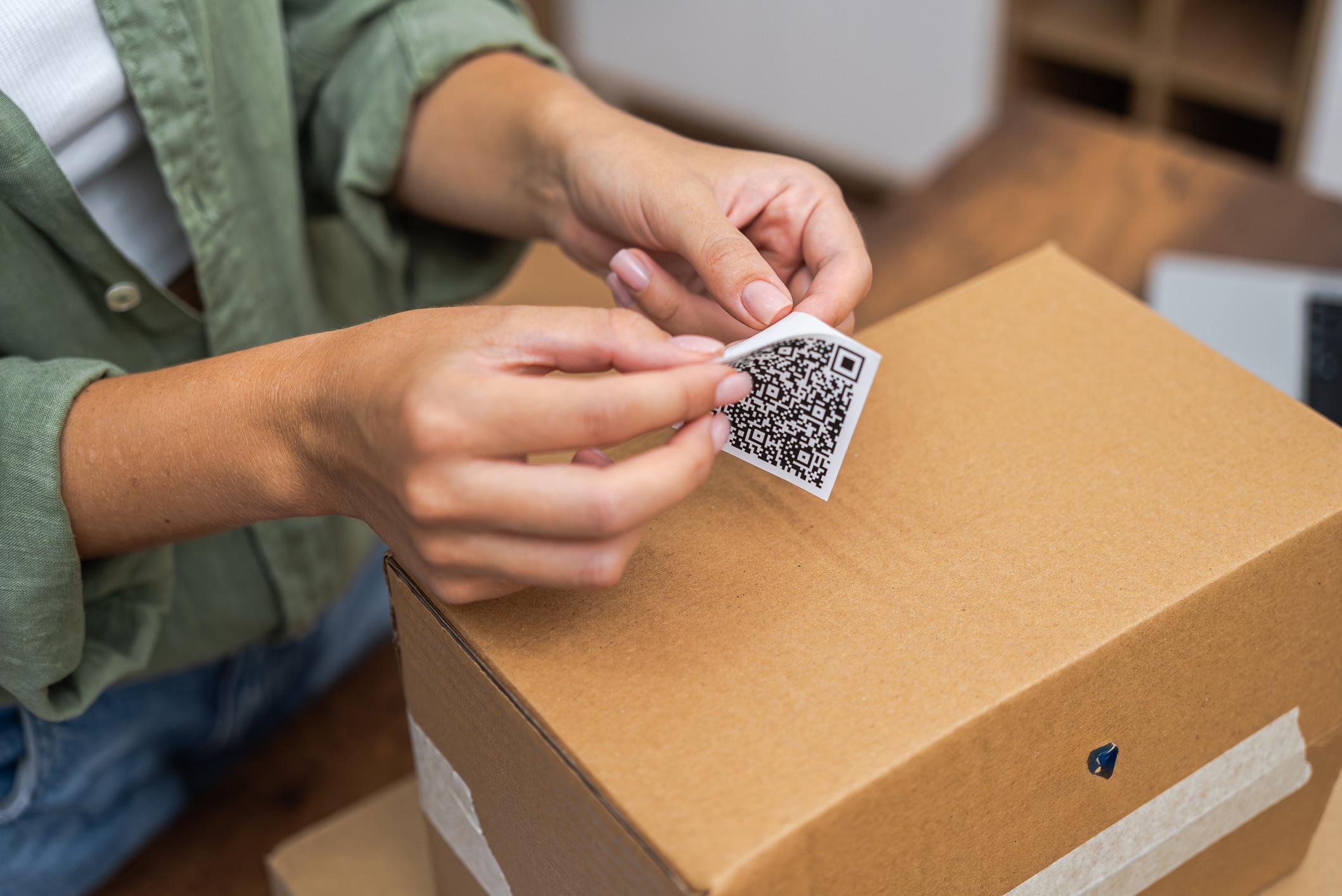 A woman's hands peeling the backing off a QR code sticker and applying it to a cardboard box on a table. She is wearing a white top, green shirt, and blue jeans, with a laptop visible on the table in the background.